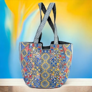 Könnte beinhalten: Eine graue Tragetasche mit einem geometrischen Muster in Rot, Blau, Gelb und Silber. Die Tasche hat zwei graue Griffe und ist mit kleinen Spiegeln verziert. Der Hintergrund ist ein Farbverlauf von Gelb und Blau.