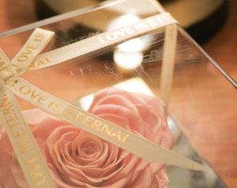 May include: A preserved pink rose in a clear, geometric glass box. The box is tied with cream-colored ribbons printed with "LOVE IS ETERNAL" in gold lettering. The background includes a blurred lamp.