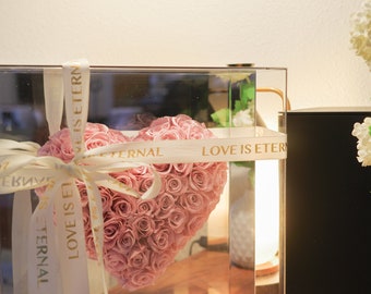 May include: A clear glass box with a pink rose heart arrangement inside. The box is tied with a white ribbon with the text "LOVE IS ETERNAL" printed in gold.