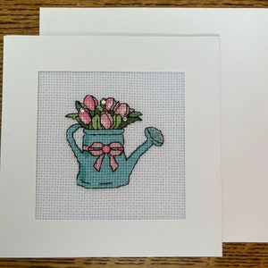 May include: A white card featuring a blue watering can filled with pink tulips. The watering can has a pink bow on the handle. The card is framed with a white border.