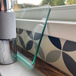 May include: A close-up of a clear glass splashback with a green silicone seal, mounted above a chrome faucet. The splashback is installed over a white countertop with a patterned tile backsplash.