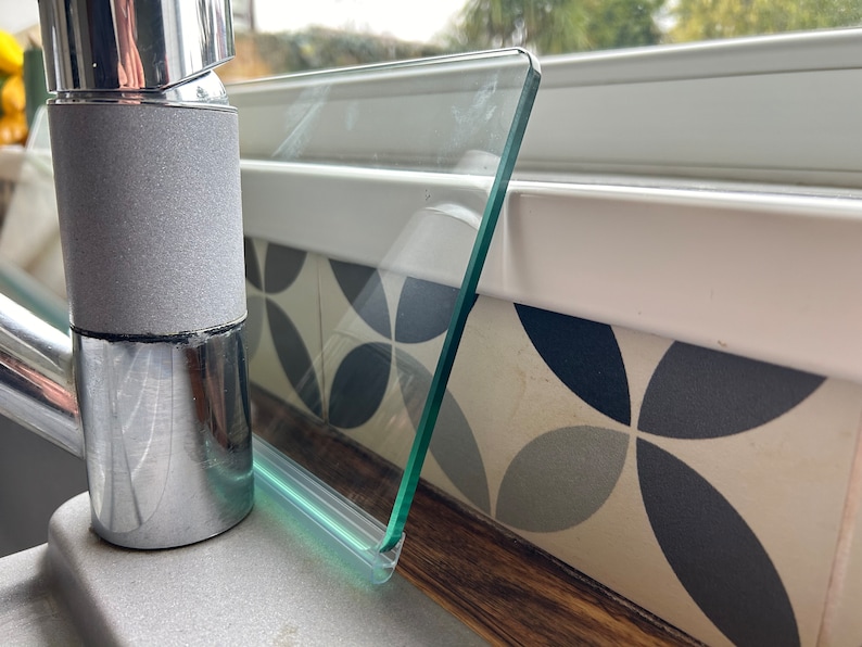 May include: A close-up of a clear glass splashback with a green silicone seal, mounted above a chrome faucet. The splashback is installed over a white countertop with a patterned tile backsplash.