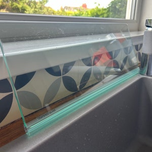 May include: A clear glass splashback with a green edge and the words "Tempered Glass" printed on it. The splashback is installed above a gray kitchen sink.