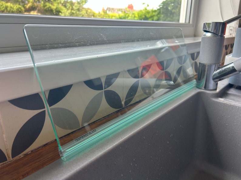 May include: A clear glass splashback with a green edge and the words "Tempered Glass" printed on it. The splashback is installed above a gray kitchen sink.
