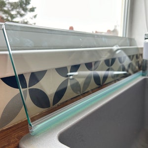 May include: A clear glass splash guard with a green trim, installed above a grey kitchen sink.