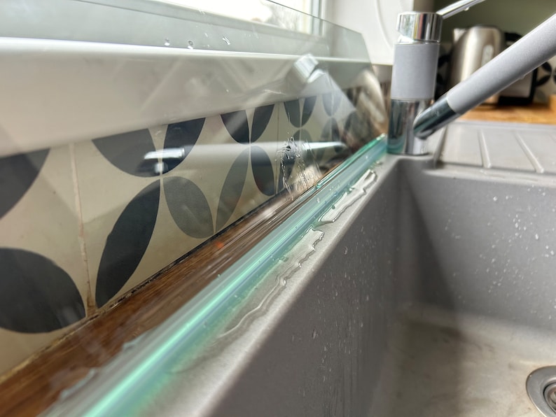 May include: A close-up of a glass splashback installed above a gray kitchen sink. The splashback is slightly angled and has a clear, reflective surface. Water droplets are visible on the surface of the splashback.