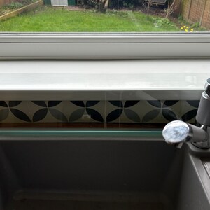 May include: A close-up of a kitchen sink with a chrome faucet. The sink is made of gray composite material and has a glass splash guard above it. The splash guard is clear and has a slight green tint. The sink is set against a backdrop of black and white patterned tile.