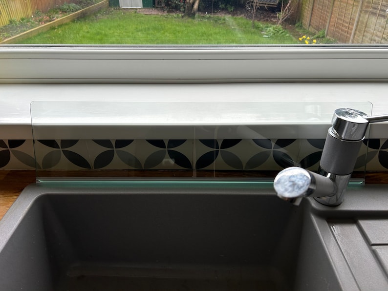 May include: A close-up of a kitchen sink with a chrome faucet. The sink is made of gray composite material and has a glass splash guard above it. The splash guard is clear and has a slight green tint. The sink is set against a backdrop of black and white patterned tile.
