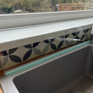 May include: A clear glass splashback installed above a gray kitchen sink. The splashback is designed to protect the wall from water splashes.