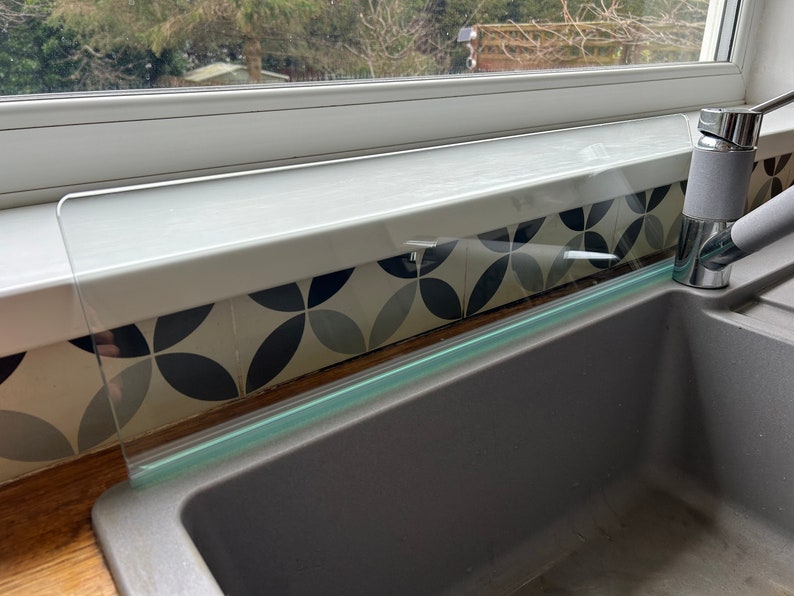 May include: A clear glass splashback installed above a gray kitchen sink. The splashback is designed to protect the wall from water splashes.