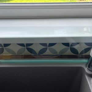 May include: A clear glass shelf with a green edge sits above a gray kitchen sink. The shelf is installed above a window with a white frame. The sink has a chrome faucet with a gray handle.