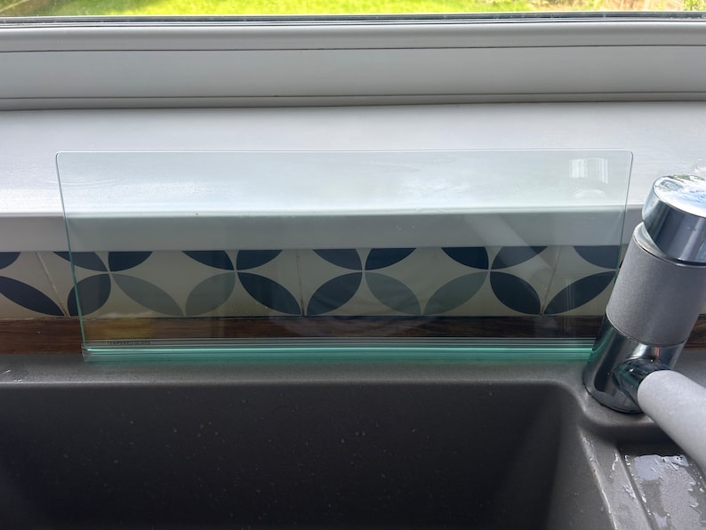May include: A clear glass shelf with a green edge sits above a gray kitchen sink. The shelf is installed above a window with a white frame. The sink has a chrome faucet with a gray handle.