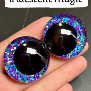 Op de afbeelding: Twee ronde, zwarte poppenogen met een glinsterende, iriserende rand van blauw, paars en roze. De tekst "Iridescent Magic" staat in een witte rechthoek boven de ogen.