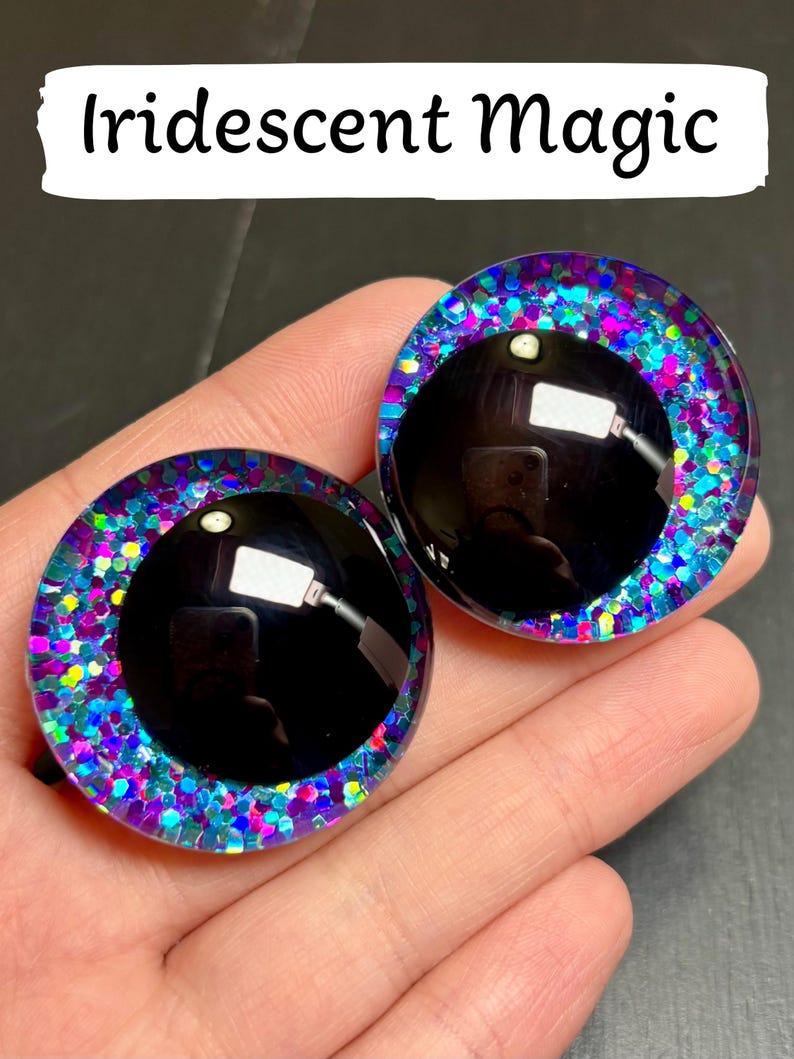 Op de afbeelding: Twee ronde, zwarte poppenogen met een glinsterende, iriserende rand van blauw, paars en roze. De tekst "Iridescent Magic" staat in een witte rechthoek boven de ogen.
