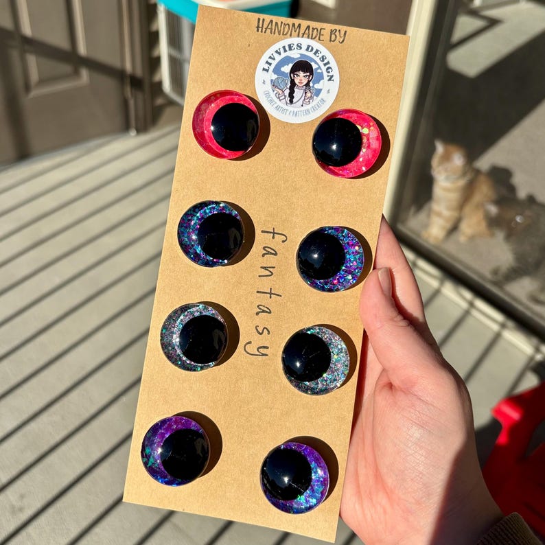 Op de afbeelding: Een verzameling handgemaakte fantasy cabochons in verschillende kleuren, waaronder roze, blauw, zilver en paars, met een zwart midden. De cabochons zijn gerangschikt op een bruine kaart met het woord "fantasy" erop gedrukt.