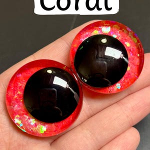 Op de afbeelding: Twee koraalkleurige, ronde knutselogen met zwarte pupillen. De ogen hebben een glinsterende, rood-oranje buitenring en zijn gelabeld met "Coral" in een witte rechthoek. Ideaal voor knutselprojecten.