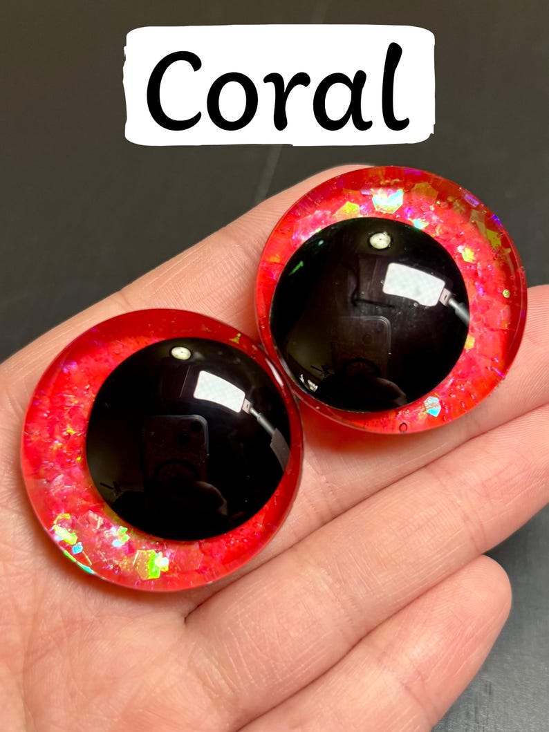 Op de afbeelding: Twee koraalkleurige, ronde knutselogen met zwarte pupillen. De ogen hebben een glinsterende, rood-oranje buitenring en zijn gelabeld met "Coral" in een witte rechthoek. Ideaal voor knutselprojecten.