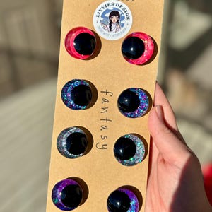 Op de afbeelding: Een set van acht handgemaakte cabochon-oogversieringen met fantasiethema. De ogen zijn zwart met kleurrijke glitteraccenten in roze, blauw en paars. Gemonteerd op een bruine kaart met de tekst "fantasy" en "handmade by Livvies Design".