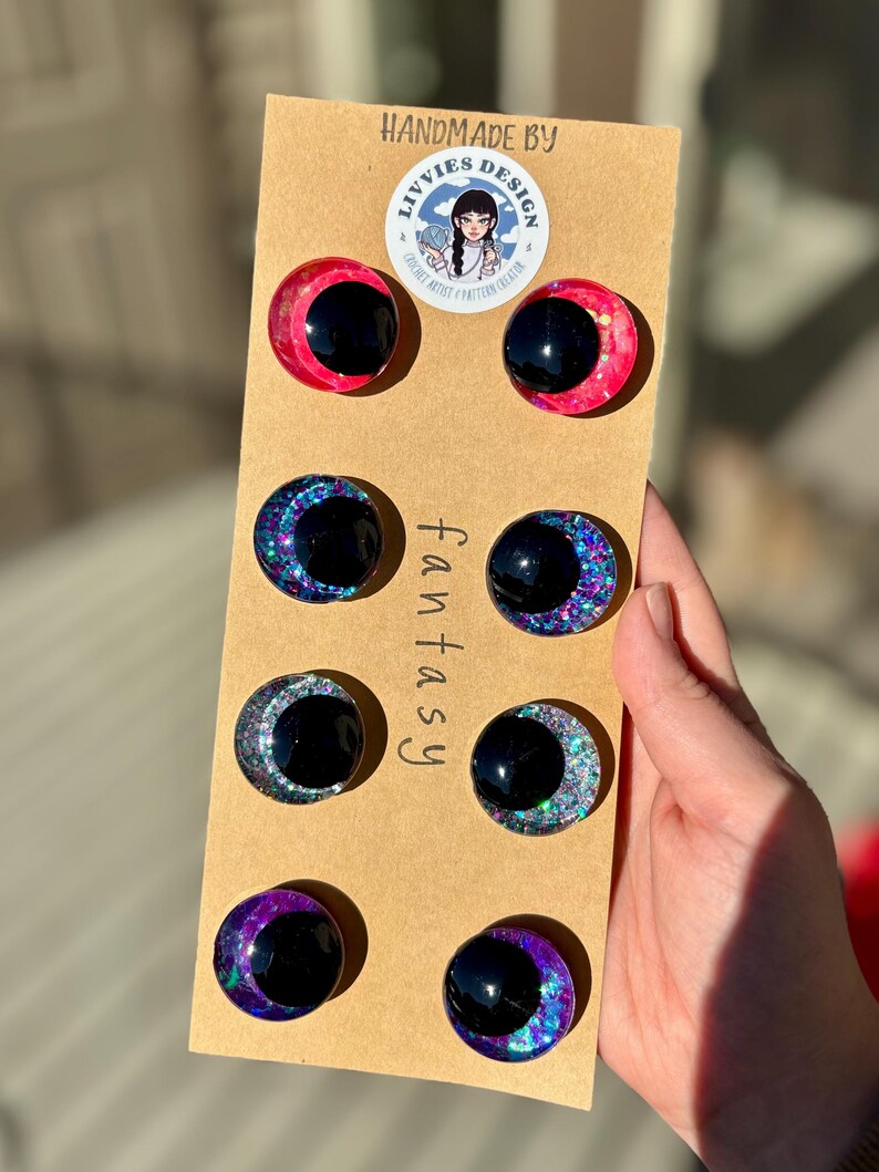 Op de afbeelding: Een set van acht handgemaakte cabochon-oogversieringen met fantasiethema. De ogen zijn zwart met kleurrijke glitteraccenten in roze, blauw en paars. Gemonteerd op een bruine kaart met de tekst "fantasy" en "handmade by Livvies Design".