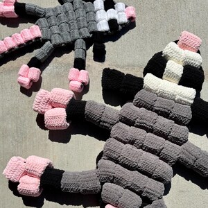 May include: Two handmade crochet dolls in a playful design. One doll features a gray body with black and white accents, while the other has a gray body with black and pink accents. The dolls have extended limbs and are made with a textured yarn.