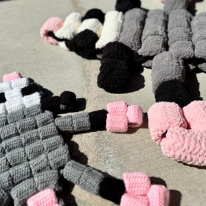 May include: Handmade crochet art in a playful design. The artwork features a gray, black, white, and pink color scheme. The pieces are arranged on a concrete surface, showcasing the texture and detail of the yarn. The design appears to be a whimsical creature.