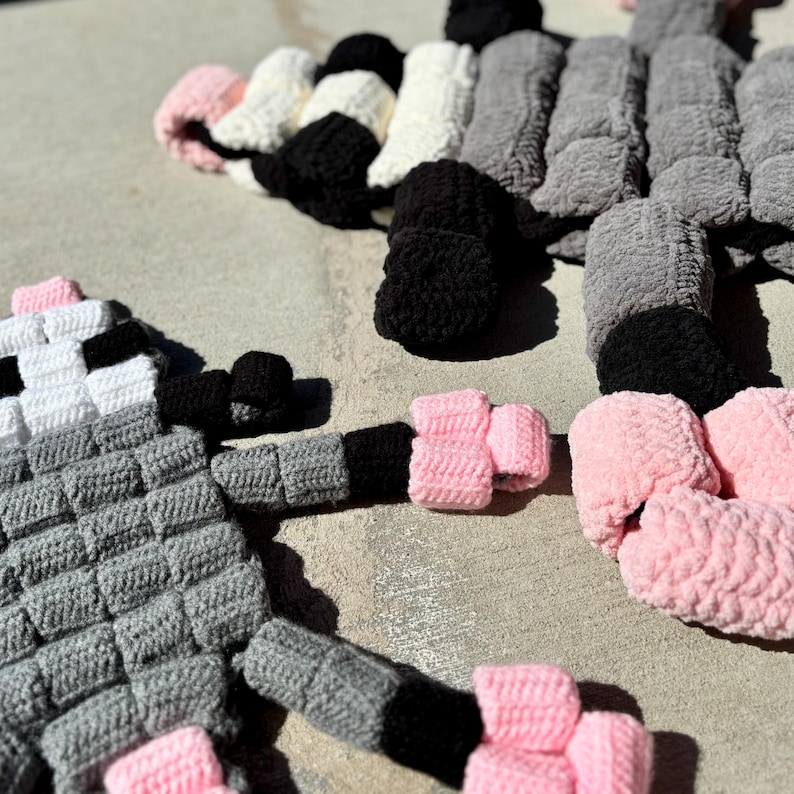 May include: Handmade crochet art in a playful design. The artwork features a gray, black, white, and pink color scheme. The pieces are arranged on a concrete surface, showcasing the texture and detail of the yarn. The design appears to be a whimsical creature.