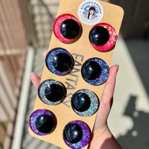 Op de afbeelding: Een set van acht fantasy-oogcabochons in verschillende kleuren, waaronder rood, blauw, groen en paars, met zwarte pupillen. De cabochons zijn gerangschikt op een bruine kartonnen achterkant met het woord "FANTASY" erop gedrukt. Het merklogo "LIVVIES DESIGN" is ook zichtbaar.