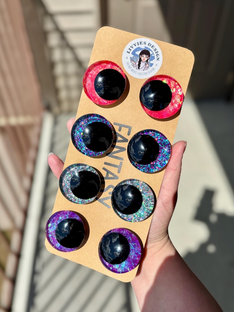 Op de afbeelding: Een set van acht fantasy-oogcabochons in verschillende kleuren, waaronder rood, blauw, groen en paars, met zwarte pupillen. De cabochons zijn gerangschikt op een bruine kartonnen achterkant met het woord "FANTASY" erop gedrukt. Het merklogo "LIVVIES DESIGN" is ook zichtbaar.