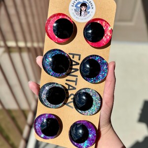 Op de afbeelding: Een set van acht fantasy-thema hars oog cabochons in verschillende kleuren, waaronder rood, blauw en paars, met glitter accenten. De ogen hebben een zwarte pupil en zijn gemonteerd op een bruine kartonnen achterkant. Het merklogo is zichtbaar.