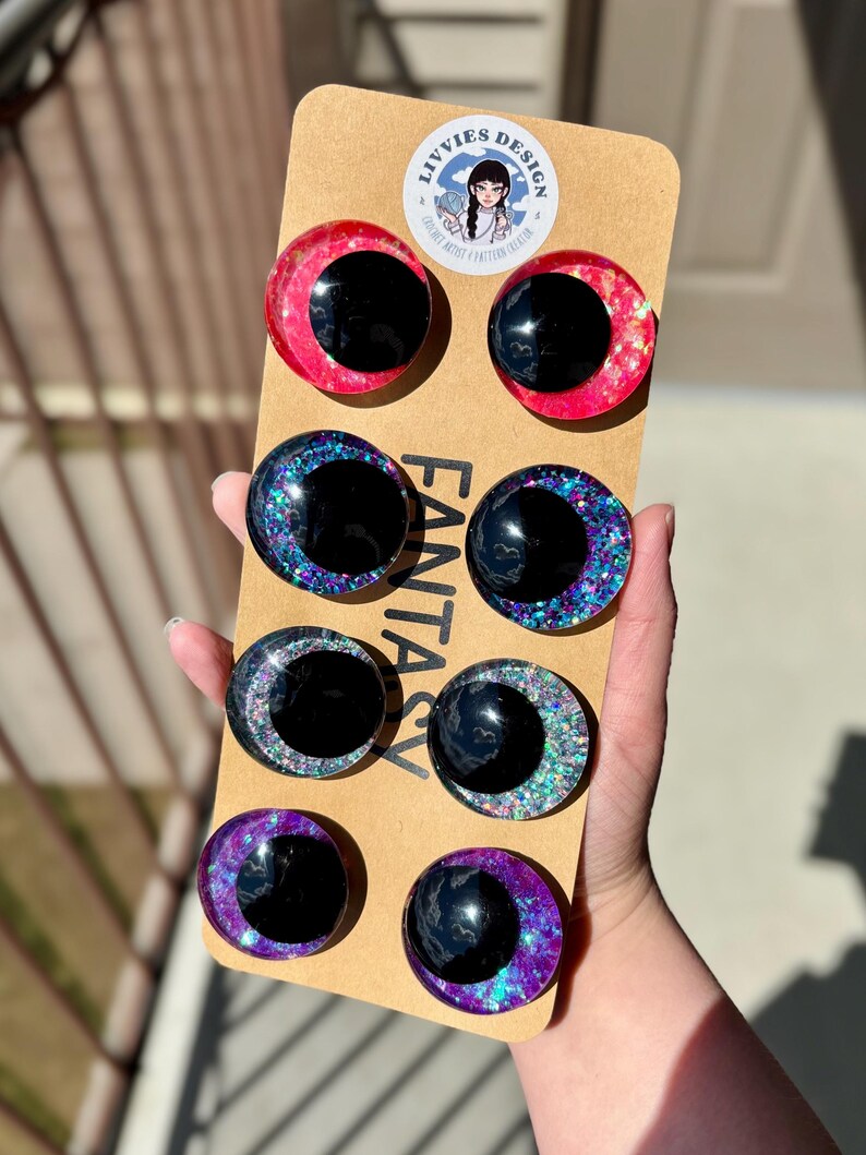 Op de afbeelding: Een set van acht fantasy-thema hars oog cabochons in verschillende kleuren, waaronder rood, blauw en paars, met glitter accenten. De ogen hebben een zwarte pupil en zijn gemonteerd op een bruine kartonnen achterkant. Het merklogo is zichtbaar.
