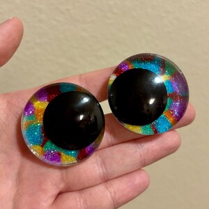 May include: Two large, round, glittery craft eyes with black pupils. The irises are a mix of vibrant colors including blue, purple, yellow, and red. The eyes are held in a hand against a neutral background.