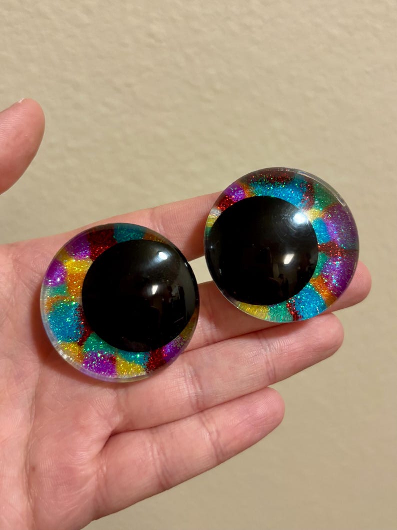 May include: Two large, round, glittery craft eyes with black pupils. The irises are a mix of vibrant colors including blue, purple, yellow, and red. The eyes are held in a hand against a neutral background.