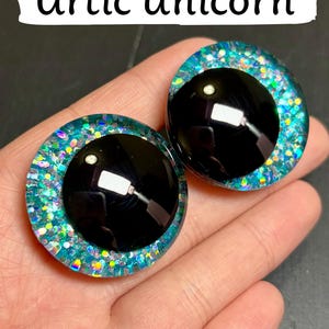 Op de afbeelding: Twee grote, ronde, zwarte poppenogen met een glinsterende, blauwgroene en iriserende rand. De tekst "Artic Unicorn" wordt weergegeven in een witte banner boven de ogen. De ogen staan tegen een donkere achtergrond.