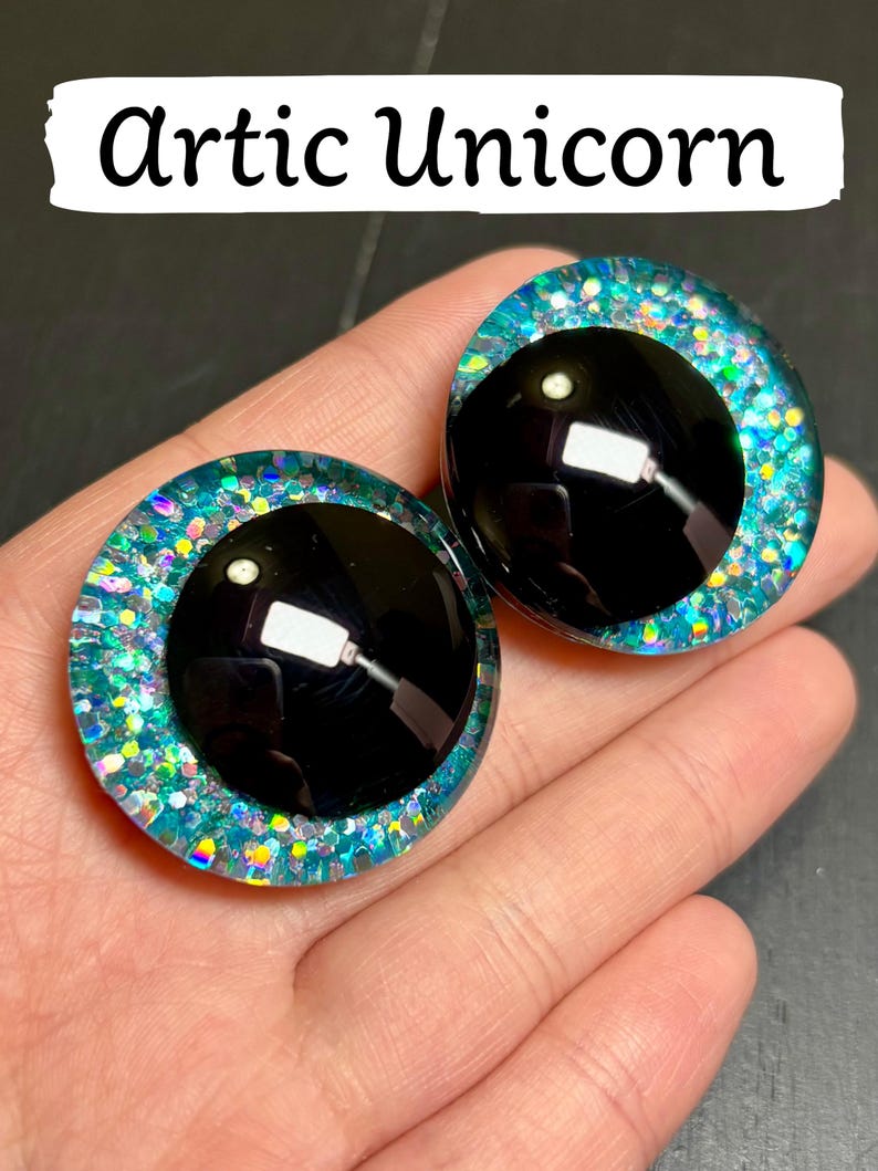 Op de afbeelding: Twee grote, ronde, zwarte poppenogen met een glinsterende, blauwgroene en iriserende rand. De tekst "Artic Unicorn" wordt weergegeven in een witte banner boven de ogen. De ogen staan tegen een donkere achtergrond.