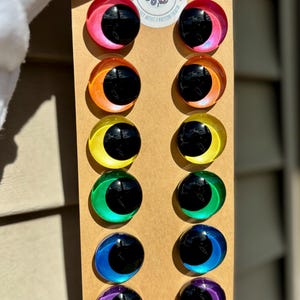 May include: A set of twelve colorful, iridescent eye-shaped craft embellishments. Each eye has a black pupil and a surrounding crescent in shades of pink, orange, yellow, green, blue, and purple. The eyes are mounted on a brown cardboard backing.