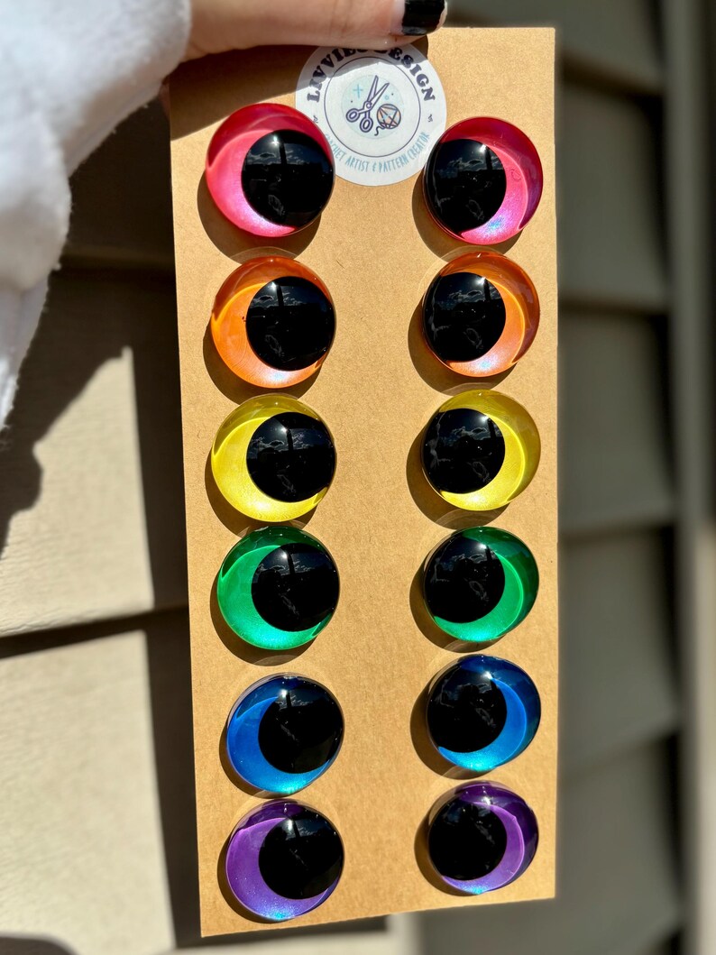 May include: A set of twelve colorful, iridescent eye-shaped craft embellishments. Each eye has a black pupil and a surrounding crescent in shades of pink, orange, yellow, green, blue, and purple. The eyes are mounted on a brown cardboard backing.