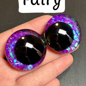 Op de afbeelding: Twee grote, ronde knutselogen met een fee&euml;nthema. De ogen hebben een zwarte pupil, omgeven door een levendige paarse en iriserende glitteriris. Het woord "Fairy" staat in zwart script boven de ogen.