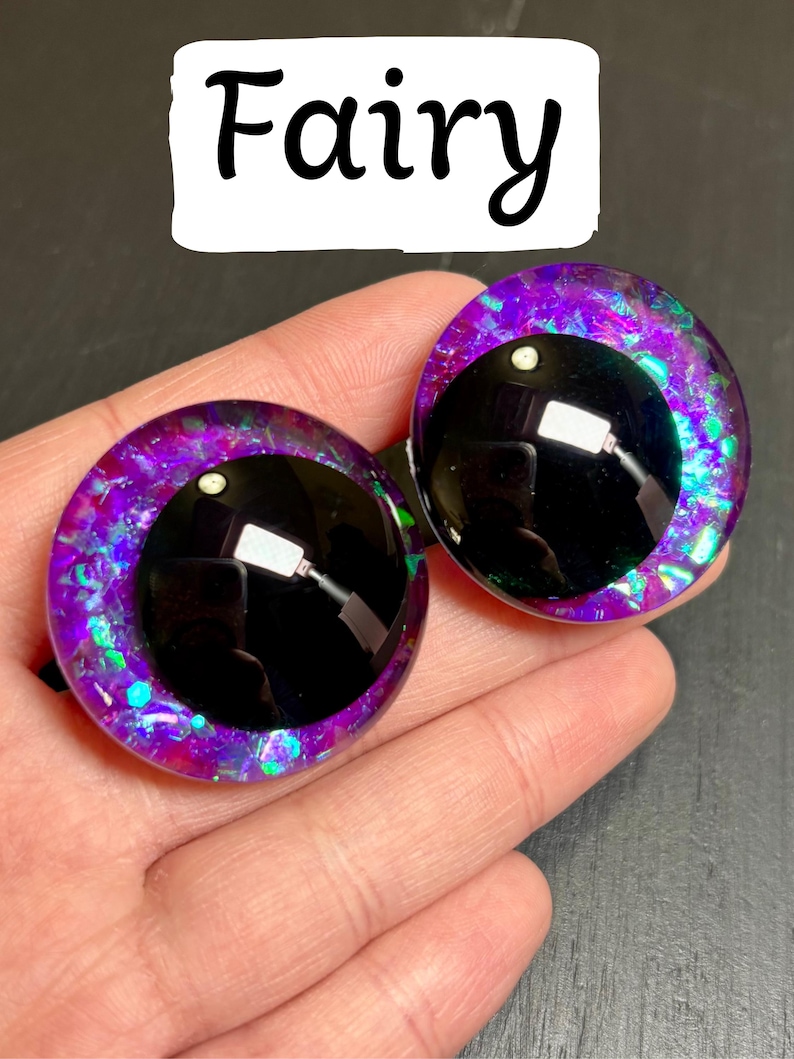 Op de afbeelding: Twee grote, ronde knutselogen met een fee&euml;nthema. De ogen hebben een zwarte pupil, omgeven door een levendige paarse en iriserende glitteriris. Het woord "Fairy" staat in zwart script boven de ogen.