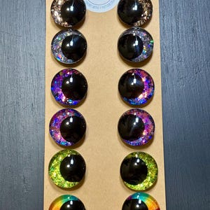 May include: A collection of twelve round, glossy, black-pupiled craft eyes with colorful, glittery crescent moon designs. The eyes are arranged in rows on a tan cardboard backing. The top of the backing features a white sticker with the text "Ivvies Design".