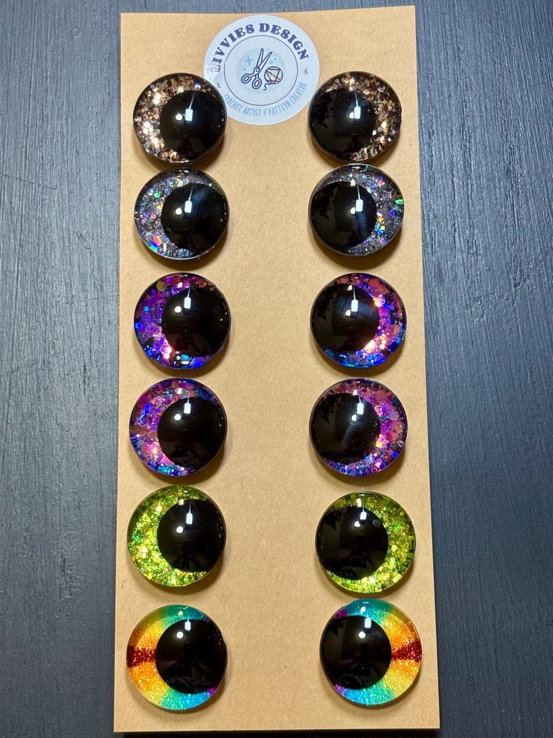 May include: A collection of twelve round, glossy, black-pupiled craft eyes with colorful, glittery crescent moon designs. The eyes are arranged in rows on a tan cardboard backing. The top of the backing features a white sticker with the text "Ivvies Design".