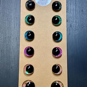 May include: A set of twelve black, round, doll eyes with glittery, colorful rims. The eyes are arranged in pairs on a brown cardboard backing. The rims feature various colors, including black, green, blue, pink, and a mix of colors. The top of the backing has a sticker with the text "Eyes Design".