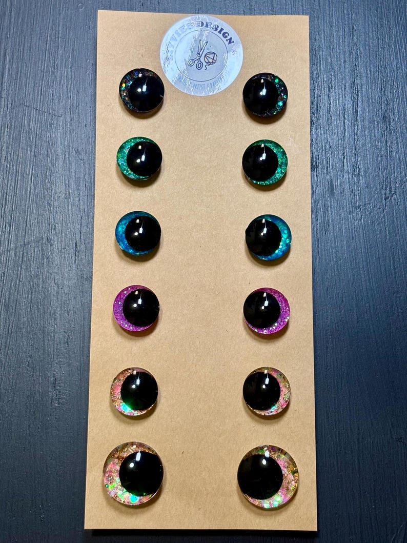 May include: A set of twelve black, round, doll eyes with glittery, colorful rims. The eyes are arranged in pairs on a brown cardboard backing. The rims feature various colors, including black, green, blue, pink, and a mix of colors. The top of the backing has a sticker with the text "Eyes Design".
