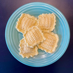May include: Five crocheted ravioli on a teal blue plate. The ravioli are yellow and have a textured surface.
