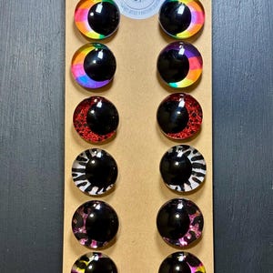 May include: A set of twelve round, decorative eye-shaped cabochons in various designs. The cabochons feature a black pupil and colorful iris patterns, including rainbow, red glitter, zebra print, and leopard print. The cabochons are arranged on a brown card.