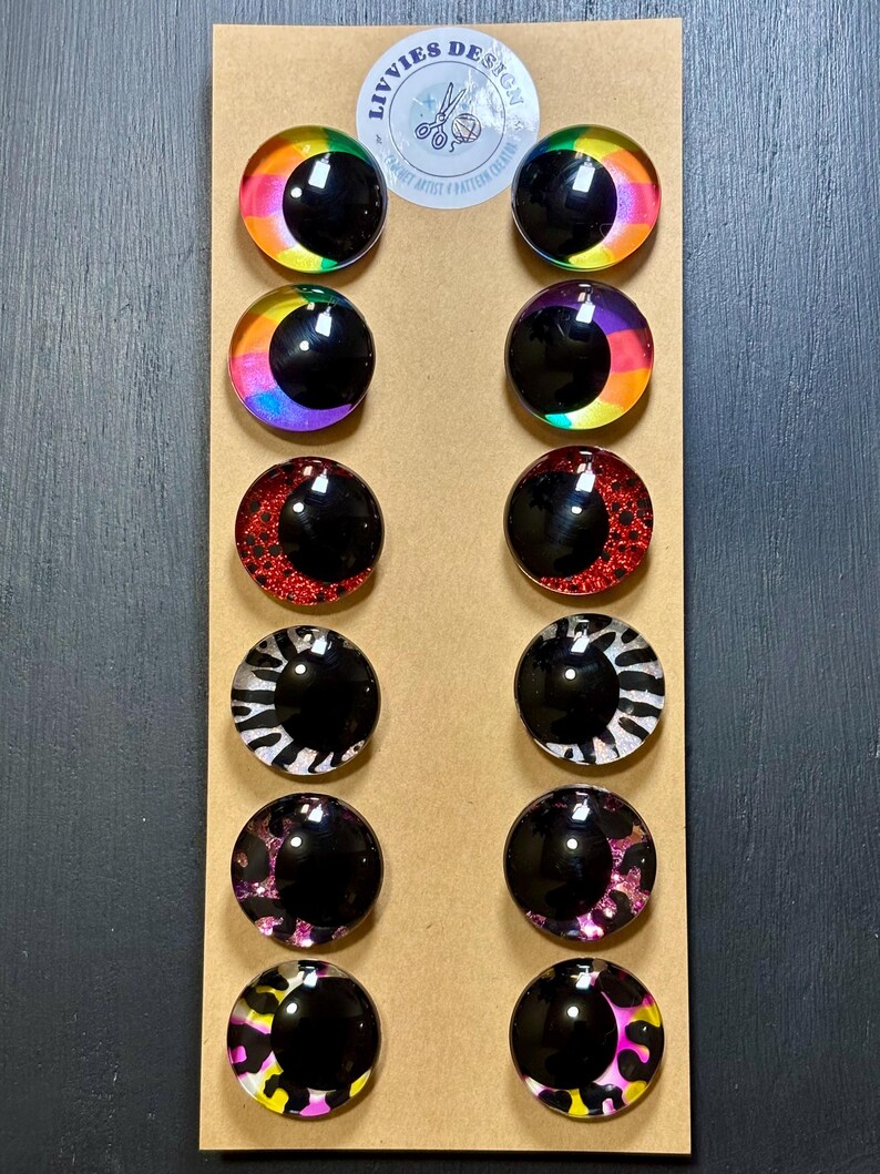 May include: A set of twelve round, decorative eye-shaped cabochons in various designs. The cabochons feature a black pupil and colorful iris patterns, including rainbow, red glitter, zebra print, and leopard print. The cabochons are arranged on a brown card.