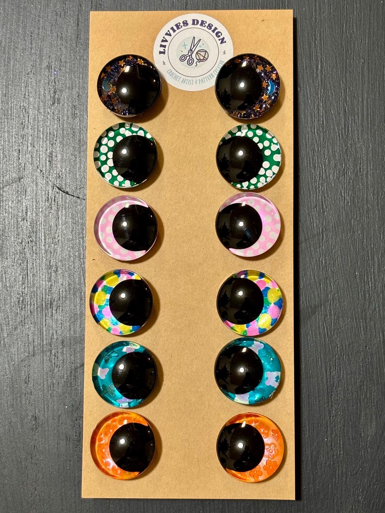 May include: A set of twelve glass cabochon eye designs in various colors and patterns, including starry night, green polka dots, pink crescent moon, and colorful abstract designs. The eyes are mounted on a brown card with the text "LIVVIES design".