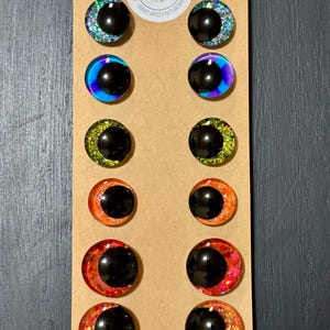May include: A collection of twelve black-centered, glitter-rimmed craft eyes in various colors, including blue, purple, green, orange, and red. The eyes are arranged on a brown cardboard backing, with a circular logo at the top that reads "LIVVIES DESIGN".