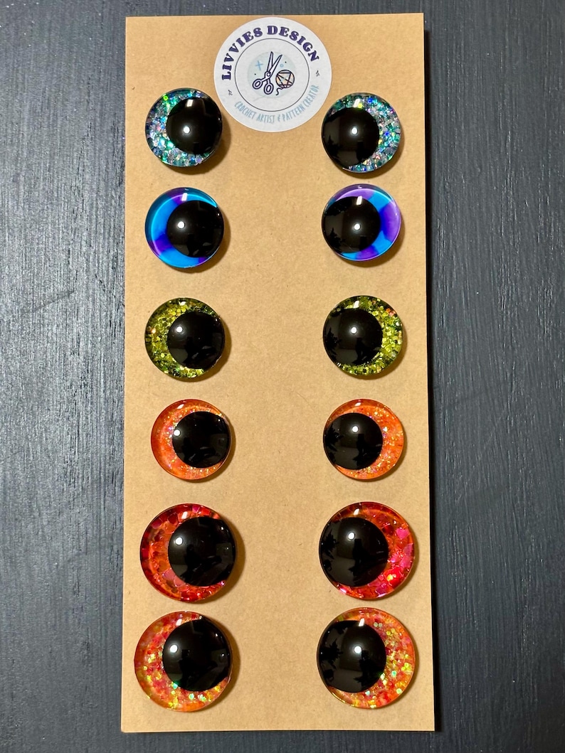 May include: A collection of twelve black-centered, glitter-rimmed craft eyes in various colors, including blue, purple, green, orange, and red. The eyes are arranged on a brown cardboard backing, with a circular logo at the top that reads "LIVVIES DESIGN".