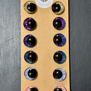 May include: A set of twelve handmade stud earrings with a crescent moon design. Each earring features a black center and a glittery, colorful border in gold, purple, blue, and pink. The earrings are displayed on a brown card with the text "LIVVIES DESIGN".