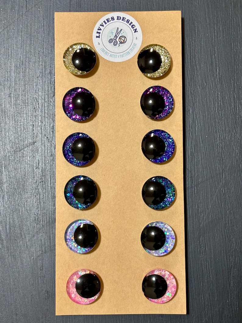 May include: A set of twelve handmade stud earrings with a crescent moon design. Each earring features a black center and a glittery, colorful border in gold, purple, blue, and pink. The earrings are displayed on a brown card with the text "LIVVIES DESIGN".